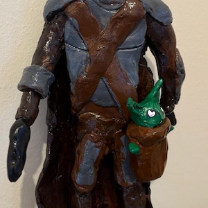 May include: A handcrafted figurine of a character in a helmet and armor, with a brown cape and a small green creature in a pouch. The figure is painted in shades of brown, gray, and black, with a glossy finish. The figure is a representation of a character from a popular science fiction series.