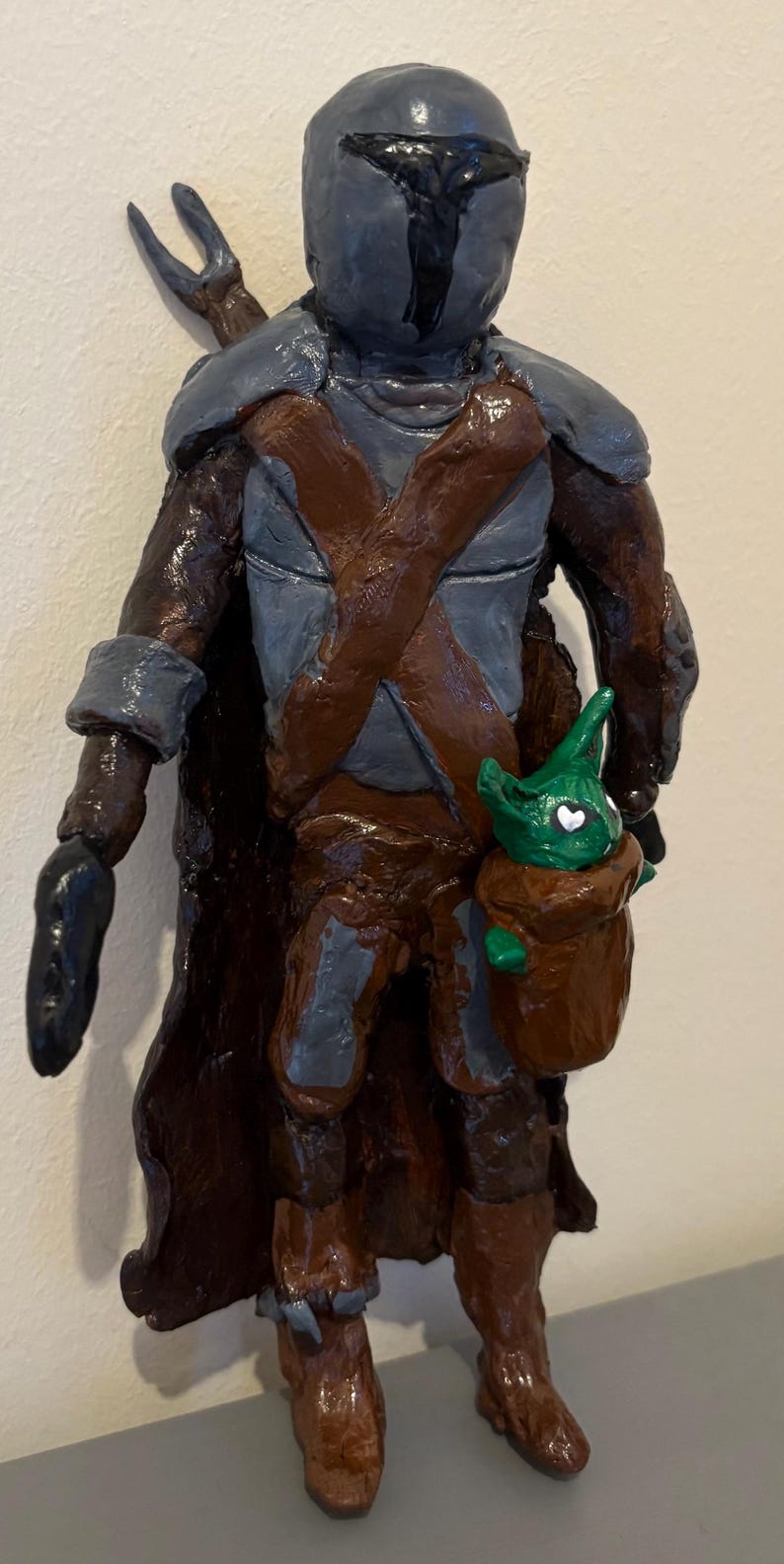 May include: A handcrafted figurine of a character in a helmet and armor, with a brown cape and a small green creature in a pouch. The figure is painted in shades of brown, gray, and black, with a glossy finish. The figure is a representation of a character from a popular science fiction series.