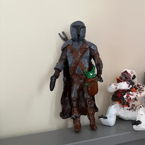 May include: A handmade figurine of a character in a helmet and armor, with a brown cloak and a small green creature in a pouch. The figure is standing, with a blaster in one hand. Another figure is seated nearby.