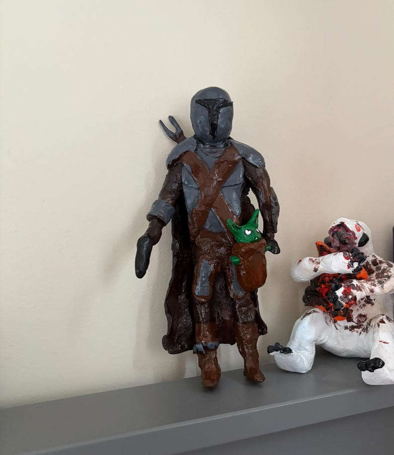 May include: A handmade figurine of a character in a helmet and armor, with a brown cloak and a small green creature in a pouch. The figure is standing, with a blaster in one hand. Another figure is seated nearby.