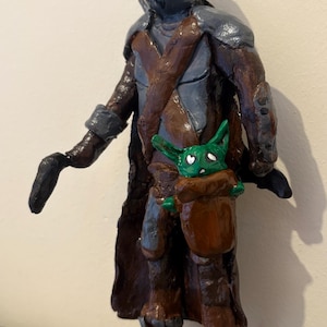 May include: A handcrafted figurine of a character in a helmet and armor, with a brown cloak and a baby-like creature in a pouch. The figure is painted in shades of gray, brown, and green.