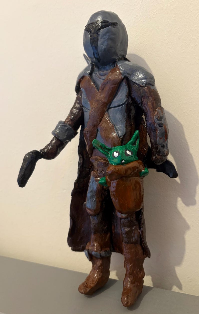 May include: A handcrafted figurine of a character in a helmet and armor, with a brown cloak and a baby-like creature in a pouch. The figure is painted in shades of gray, brown, and green.