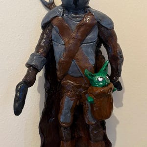 May include: A handcrafted figurine of a character in dark armor with a helmet, holding a small green creature in a brown pouch. The figure has a brown cape and boots, with gray and brown accents. The small creature has heart-shaped eyes.