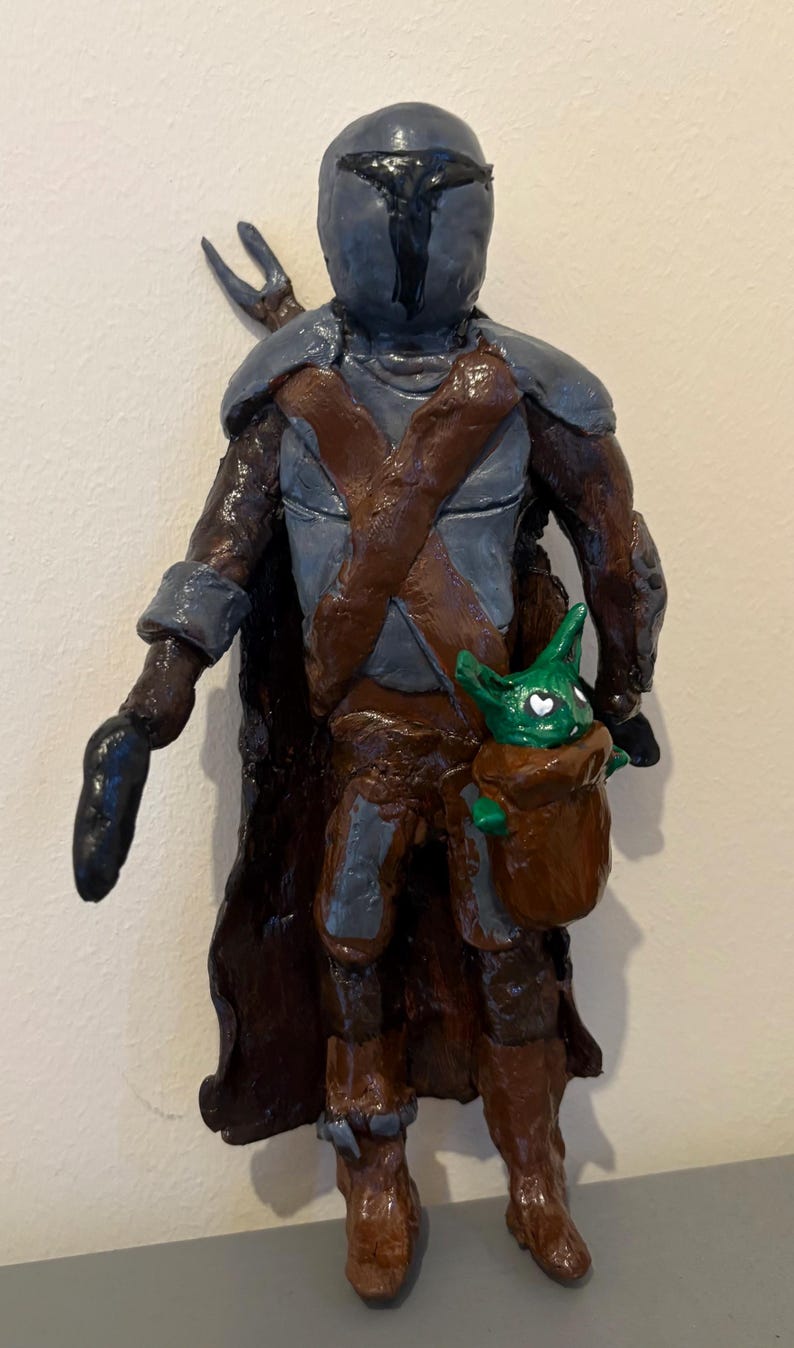 May include: A handcrafted figurine of a character in dark armor with a helmet, holding a small green creature in a brown pouch. The figure has a brown cape and boots, with gray and brown accents. The small creature has heart-shaped eyes.
