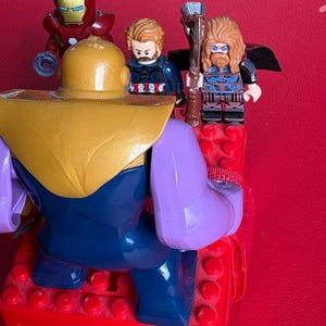 May include: A large purple and gold Thanos figure stands facing three smaller minifigures: a red and gold Iron Man, a brown-haired Captain America, and a long-haired Thor holding a brown axe, all positioned on a red plastic building block base.