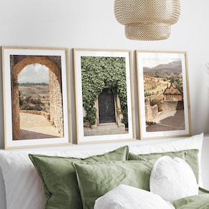 May include: Three framed landscape prints with light wood frames. The prints depict scenes of stone archways, a door covered in ivy, and a rural village. The prints are displayed above a bed with green cushions and white bedding.