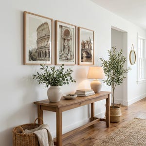 May include: A wooden console table holds a ceramic vase with green foliage, a stack of books, and a beige lamp. Three framed prints of Roman architecture hang above the table. A woven basket sits on the floor, and a potted olive tree stands nearby.