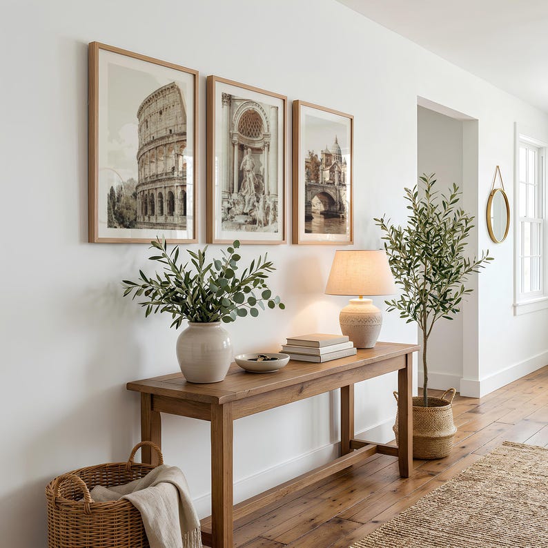 May include: A wooden console table holds a ceramic vase with green foliage, a stack of books, and a beige lamp. Three framed prints of Roman architecture hang above the table. A woven basket sits on the floor, and a potted olive tree stands nearby.