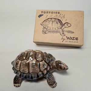 Peut inclure: Une figurine de tortue en céramique avec une carapace marron et beige, des détails bleus et une finition brillante. La figurine est présentée avec sa boîte en carton d'origine, qui porte le mot "TORTOISE" et une illustration de tortue. La boîte indique également "by WADE of ENGLAND."