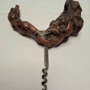 Può includere: Un cavatappi rustico con un manico in legno naturale. Il manico è di colore marrone scuro con una forma unica e strutturata. Il cavatappi in metallo è argentato e ha un design a spirale. Una piccola placca di metallo è attaccata al legno.