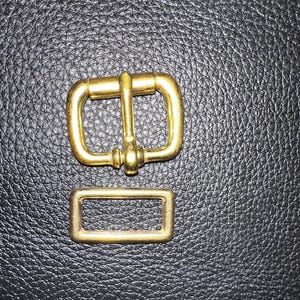 Pu&ograve; includere: Un set di fibbie in metallo color oro, composto da un telaio rettangolare e una fibbia pi&ugrave; grande con una barra centrale e un perno. Il set &egrave; esposto su una superficie nera strutturata, probabilmente in pelle o materiale simile.