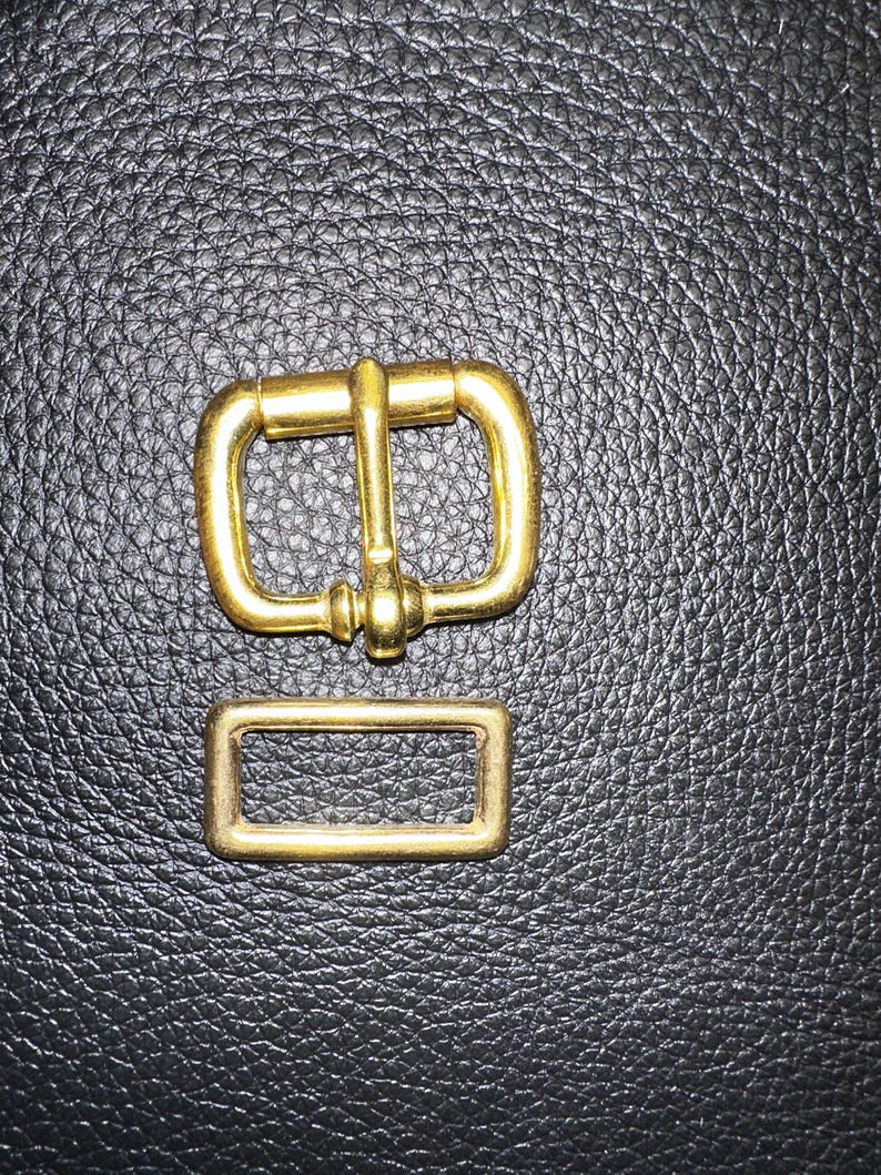 Pu&ograve; includere: Un set di fibbie in metallo color oro, composto da un telaio rettangolare e una fibbia pi&ugrave; grande con una barra centrale e un perno. Il set &egrave; esposto su una superficie nera strutturata, probabilmente in pelle o materiale simile.