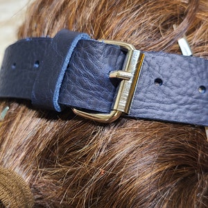 Pu&ograve; includere: Primo piano di una cintura in pelle blu navy con fibbia color argento. La cintura ha una superficie strutturata ed &egrave; fissata con una fibbia in metallo. La cintura &egrave; probabilmente per l'abbigliamento.