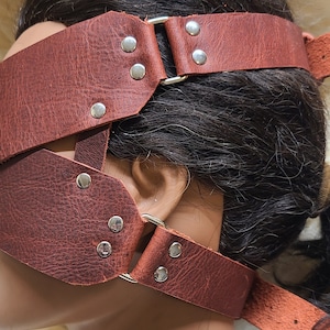 Pu&ograve; includere: Una benda per gli occhi in pelle marrone con elementi metallici argentati. La benda ha pi&ugrave; cinturini ed &egrave; fissata con rivetti e anelli di metallo. La pelle appare invecchiata.