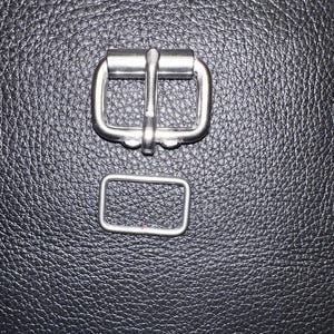 Pu&ograve; includere: Primo piano di due elementi metallici color argento. Uno &egrave; una fibbia con una barra centrale, l'altro &egrave; un anello rettangolare. L'hardware &egrave; posto su uno sfondo scuro e strutturato, probabilmente pelle o un materiale simile.
