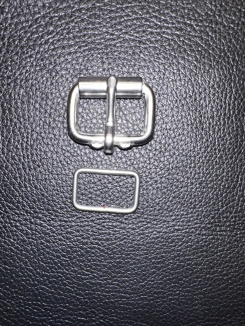 Pu&ograve; includere: Primo piano di due elementi metallici color argento. Uno &egrave; una fibbia con una barra centrale, l'altro &egrave; un anello rettangolare. L'hardware &egrave; posto su uno sfondo scuro e strutturato, probabilmente pelle o un materiale simile.
