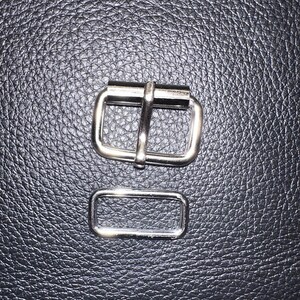 Pu&ograve; includere: Primo piano di due elementi metallici color argento. L'elemento pi&ugrave; grande &egrave; una fibbia rettangolare con una barra centrale. L'elemento pi&ugrave; piccolo &egrave; un anello rettangolare abbinato. Sfondo nero strutturato.