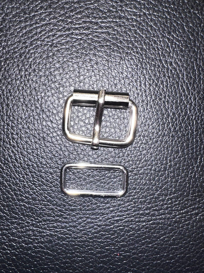 Pu&ograve; includere: Primo piano di due elementi metallici color argento. L'elemento pi&ugrave; grande &egrave; una fibbia rettangolare con una barra centrale. L'elemento pi&ugrave; piccolo &egrave; un anello rettangolare abbinato. Sfondo nero strutturato.