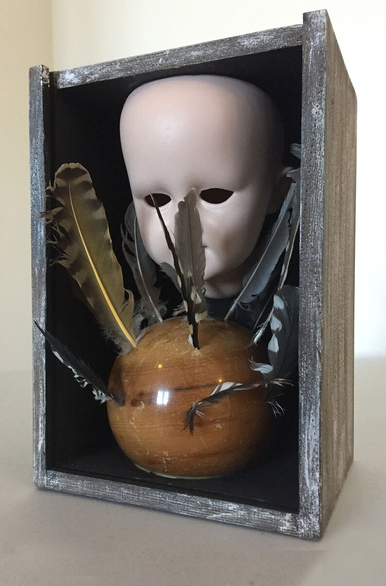 May include: A wooden box with a glass front contains a porcelain doll head, a wooden sphere, and a collection of feathers. The doll head has closed eyes and a neutral expression. The feathers are arranged around the sphere, creating a whimsical and slightly eerie display.