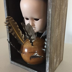May include: A wooden box with a glass front, containing a porcelain doll head, a wooden sphere, and feathers. The box is painted in a distressed gray finish.
