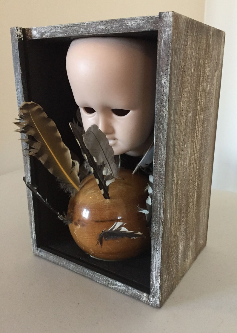 May include: A wooden box with a glass front, containing a porcelain doll head, a wooden sphere, and feathers. The box is painted in a distressed gray finish.
