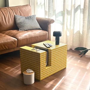 May include: A yellow tiled coffee table with a brown leather sofa in the background. The coffee table has a magazine and a small white box on top.