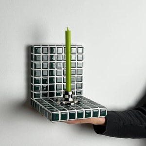 Puede incluir: Un pequeño estante de azulejos verdes y blancos con una vela verde en un candelabro blanco y negro. El estante está siendo sostenido por una mano.
