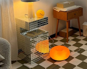 May include: A mirrored side table with a gray lamp and a yellow smiley face lamp. The table is on a black and white checkered rug. There is a wooden side table with a drawer and a box on top of it. The wall behind the tables has a framed print with the text "about me" and various icons.