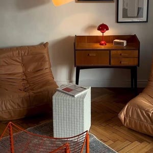 May include: An interior scene featuring a mid-century modern aesthetic. A brown wooden cabinet with drawers is topped with a red mushroom lamp. Artwork hangs on the wall above, and two brown leather chairs are on either side.