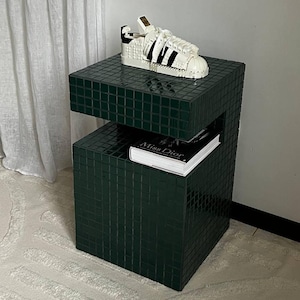 May include: A dark green tiled end table with two shelves. The top shelf has a white Lego shoe on it. The bottom shelf has a book titled "Miss Dior" on it.