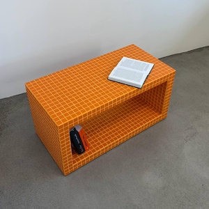 Puede incluir: Una mesa de centro rectangular naranja con un patrón de cuadrícula. Un libro descansa encima y otro es visible dentro del estante abierto. La mesa está hecha de un material sólido y se encuentra sobre un suelo gris.