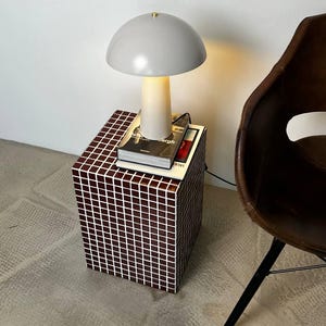 May include: A modern gray table lamp with a rounded shade and a cream-colored base. The lamp sits atop a cube-shaped side table covered in a mosaic tile pattern. Books are stacked under the lamp. A brown leather chair is in the background.