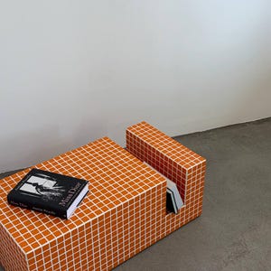 May include: An orange and white tiled coffee table with a matching side table. A book titled "Miss Dior" is on the table. The table and side table are made of small, square tiles.