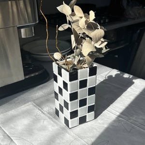 Puede incluir: Un jarrón rectangular con un patrón de tablero de ajedrez en blanco y negro. El jarrón está lleno de hojas de eucalipto secas y ramas marrones delgadas. El jarrón está sobre un mantel blanco.