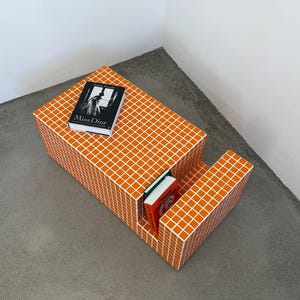 May include: An orange and white tiled coffee table with a built-in shelf. A book titled "Miss Dior" rests on top, and two books are inside the shelf. The table is a geometric design, perfect for a modern living space.