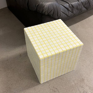 May include: A cube-shaped side table with a grid pattern of white squares and yellow grout. The table is made of small, square tiles. The table is sitting on a concrete floor, with a black leather sofa in the background.