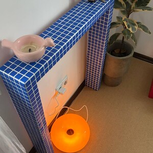 Puede incluir: Una consola con azulejos azules con un candelabro rosa en forma de pájaro y una lámpara redonda naranja. La mesa tiene un estante con una botella y llaves. Una planta en maceta está al lado de la mesa. La lámpara está en el suelo.