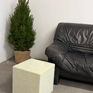May include: A cube-shaped side table with a mosaic tile design. The tiles are white with yellow grout lines. A small evergreen tree in a burlap pot and a black leather sofa are in the background.