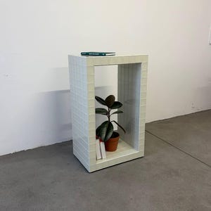 May include: A white, rectangular side table with a grid tile design. A potted plant with dark green leaves and a small book are inside the table's open space. A book rests on top of the table.