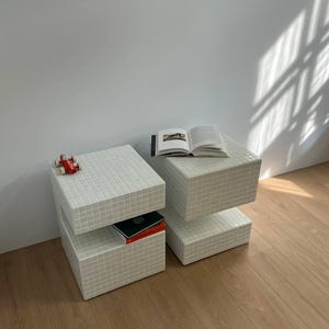 May include: Two white square end tables with a tiled surface. The table on the left has a red toy car on top and a stack of books on the lower shelf. The table on the right has a book on top.