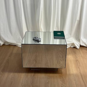Puede incluir: Una mesa de centro espejada de forma rectangular con una figura de coche plateada encima. También hay un libro verde sobre la mesa. La mesa está sobre un suelo de madera, con una cortina blanca al fondo.
