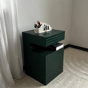 May include: A dark green square side table with a tiled surface. A white and black sneaker sits on the top shelf. A book is on the lower shelf.