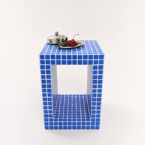 Könnte beinhalten: Ein blauer, gefliester Beistelltisch mit quadratischer Oberseite und offenen Seiten. Ein kleines, rundes, silbernes Tablett mit Deckel und ein kleiner Teller mit rotem Essen und einer Kirsche stehen darauf. Der Tisch ist ein einzigartiges, modernes Design.