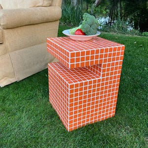 Puede incluir: Una mesa auxiliar de mosaico naranja de dos niveles con lechada blanca, colocada sobre hierba verde. Un plato de brócoli y un tomate se encuentra en el nivel superior. Un sillón beige está en el fondo.