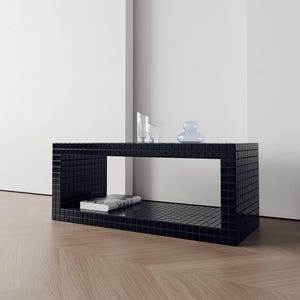 Puede incluir: Una mesa de centro rectangular negra con una superficie de azulejos. La mesa tiene un estante abierto y contiene un libro, un vaso transparente y un objeto decorativo azul claro. La mesa está sobre un suelo de madera.