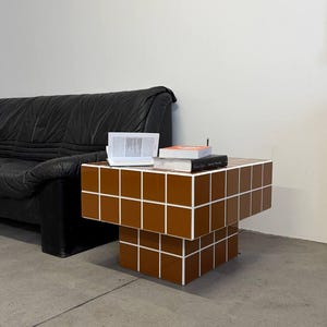 May include: A brown and white tiled side table with a stacked design. The table has a rectangular top and base, with a series of brown tiles outlined in white. Books and a notepad rest on the table's surface. A black leather sofa is in the background.