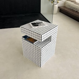 May include: A modern, cube-shaped side table with a black and white grid pattern. The table has a small shelf and holds a magazine and a book. The design is minimalist and geometric, suitable for a contemporary living space.