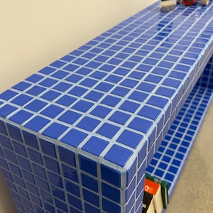 Puede incluir: Una consola azul con azulejos y una balda inferior. La mesa está decorada con un pequeño coche de juguete rojo y una fotografía enmarcada. Los azulejos son cuadrados y están dispuestos en un patrón de cuadrícula.