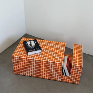 May include: An orange and white grid-patterned coffee table with a cutout section. The table holds two books, one black with the title "Miss Dior" and another with a red cover. The table is placed in a corner against a white wall.