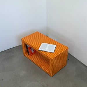 Puede incluir: Una mesa de centro rectangular naranja con un patrón de cuadrícula y un estante. Un libro abierto está encima y una pila de libros está dentro del estante. La mesa está en una esquina de una habitación con paredes blancas y suelo gris.